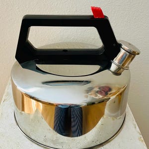 May include: A stainless steel stovetop kettle with a black handle and a red lever on the spout. The kettle has a shiny, reflective surface and a rounded base. The handle is rectangular and angled.