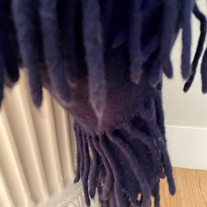 May include: Close-up of a navy blue fringed throw blanket. The blanket's fringe hangs down, revealing the texture and colour of the fabric. The throw is draped over a white decorative urn, with a wooden floor in the background.
