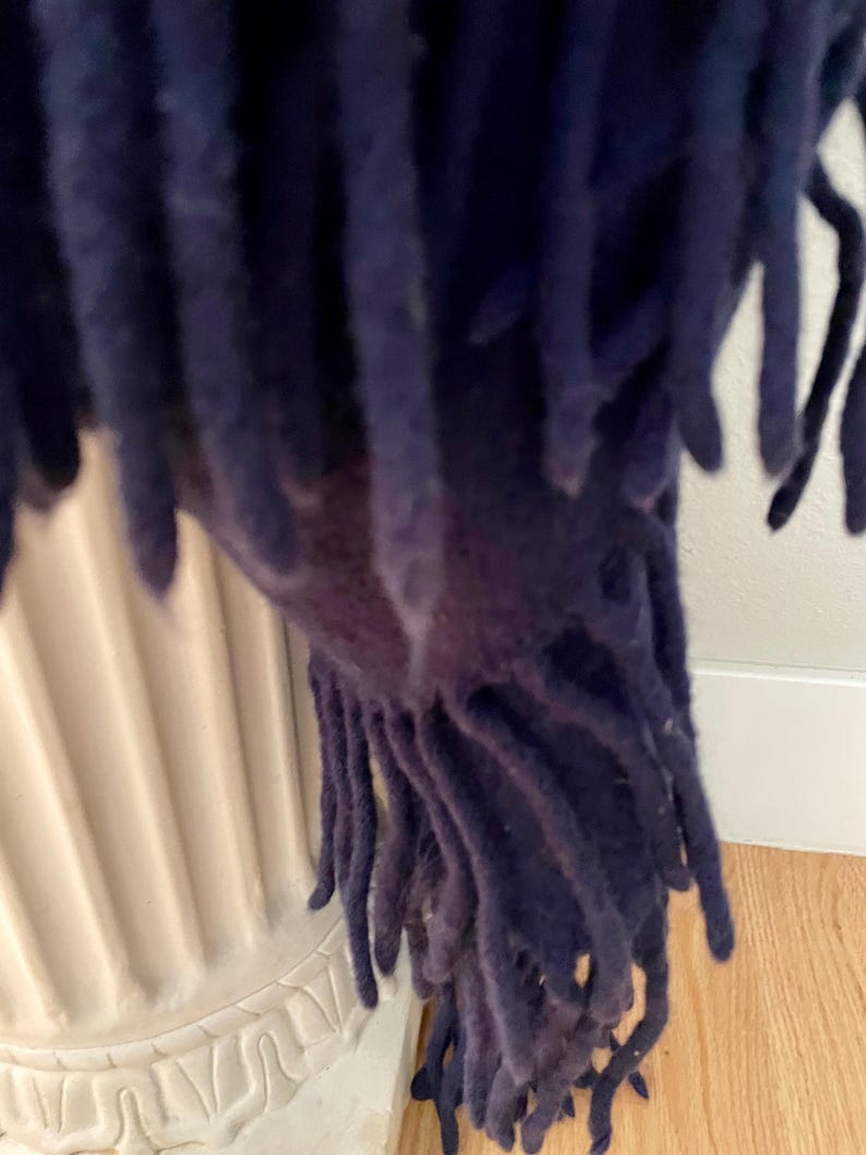 May include: Close-up of a navy blue fringed throw blanket. The blanket's fringe hangs down, revealing the texture and colour of the fabric. The throw is draped over a white decorative urn, with a wooden floor in the background.