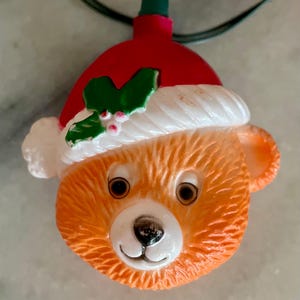 May include: A Christmas light shaped like a teddy bear's head. The bear is orange with a white and red Santa hat. The hat has a sprig of holly. The light has a green cord.