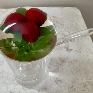 May include: A clear glass cup with a handle, containing red raspberries and green leaves encased in a clear, rounded top. The cup sits on a white surface.
