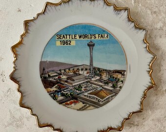 May include: A white ceramic plate with a gold rim and a colorful illustration of the Seattle World's Fair in 1962. The illustration includes the Space Needle and other buildings.