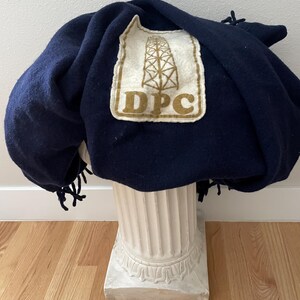May include: A navy blue blanket with a cream-coloured patch featuring a brown oil derrick and the letters "DPC". The blanket has a fringe along the edge and is draped over a white pedestal.