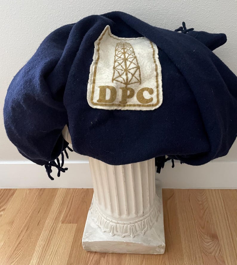May include: A navy blue blanket with a cream-coloured patch featuring a brown oil derrick and the letters "DPC". The blanket has a fringe along the edge and is draped over a white pedestal.