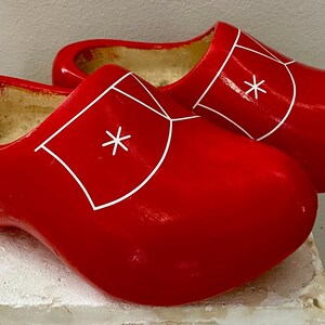 May include: A pair of bright red wooden clogs. Each clog features a white geometric design with a star-shaped accent. The clogs are displayed on a light-colored surface, showcasing their vibrant color and traditional design.