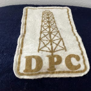 May include: A white felt patch featuring a brown oil derrick design and the letters "DPC". The patch has a brown border and is set against a dark blue background. The patch is rectangular with rounded corners.