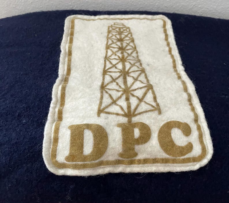 May include: A white felt patch featuring a brown oil derrick design and the letters "DPC". The patch has a brown border and is set against a dark blue background. The patch is rectangular with rounded corners.