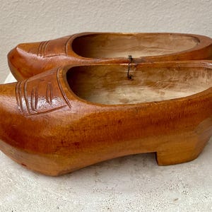 May include: A pair of wooden clogs, showcasing a warm, medium-brown hue. The clogs have a classic design with a rounded toe and a small heel. Decorative grooves are carved into the top of each shoe. The clogs are displayed on a light-colored surface.