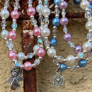 Könnte beinhalten: Mehrere Armbänder mit perlenartigen Perlen in Rosa, Weiß und Blau. Klare Kristallperlen und silberne Charms, darunter ein Engel und ein religiöses Emblem. Weiße Kreuz-Charms sind ebenfalls vorhanden.