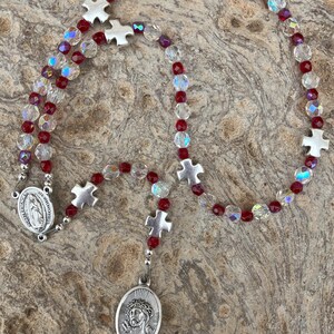 K&ouml;nnte beinhalten: Ein Rosenkranz mit roten und klaren Kristallperlen und silberfarbenen Metallkreuz- und ovalen Anh&auml;ngern. Der Anh&auml;nger zeigt ein Bild von Jesus. Der Rosenkranz wird auf einer strukturierten, neutral get&ouml;nten Oberfl&auml;che pr&auml;sentiert.