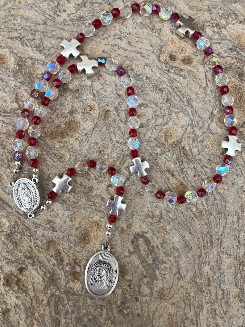 K&ouml;nnte beinhalten: Ein Rosenkranz mit roten und klaren Kristallperlen und silberfarbenen Metallkreuz- und ovalen Anh&auml;ngern. Der Anh&auml;nger zeigt ein Bild von Jesus. Der Rosenkranz wird auf einer strukturierten, neutral get&ouml;nten Oberfl&auml;che pr&auml;sentiert.
