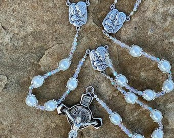 Puede incluir: Un rosario con grandes perlas blancas y detalles metálicos plateados. El rosario presenta un crucifijo con una detallada representación de Jesús y varios medallones decorativos. Las cuentas están ensartadas en un delicado alambre transparente.