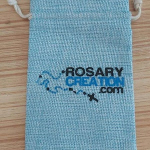 K&ouml;nnte beinhalten: Hellblaues Beutel mit Kordelzug und dem Text "ROSARY CREATION .com" in Schwarz und Blau. Der Beutel hat eine strukturierte Oberfl&auml;che und einen wei&szlig;en Kordelverschluss.