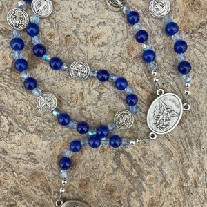 May include: A blue and clear crystal rosary with silver medallions featuring religious imagery. The medallions depict Saint Michael and Saint Anthony.