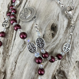 K&ouml;nnte beinhalten: Ein Rosenkranz mit weinroten Perlen und klaren Kristallakzenten. Der Rosenkranz hat silberfarbene Metallkomponenten, darunter ein Kruzifix, ovale kreuzf&ouml;rmige Perlen und ein Medaillon mit einem religi&ouml;sen Bild.