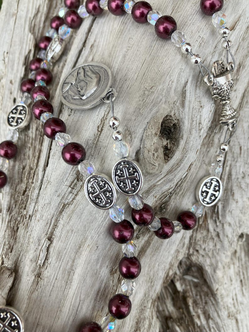 K&ouml;nnte beinhalten: Ein Rosenkranz mit weinroten Perlen und klaren Kristallakzenten. Der Rosenkranz hat silberfarbene Metallkomponenten, darunter ein Kruzifix, ovale kreuzf&ouml;rmige Perlen und ein Medaillon mit einem religi&ouml;sen Bild.