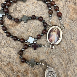 K&ouml;nnte beinhalten: Ein Rosenkranz mit dunkelroten und grauen Perlen, durchsetzt mit silberfarbenen Perlen und kreuzf&ouml;rmigen Akzenten. Der Rosenkranz hat zwei ovale Anh&auml;nger, einen mit einem Bild von Jesus und den anderen mit einer religi&ouml;sen Figur.