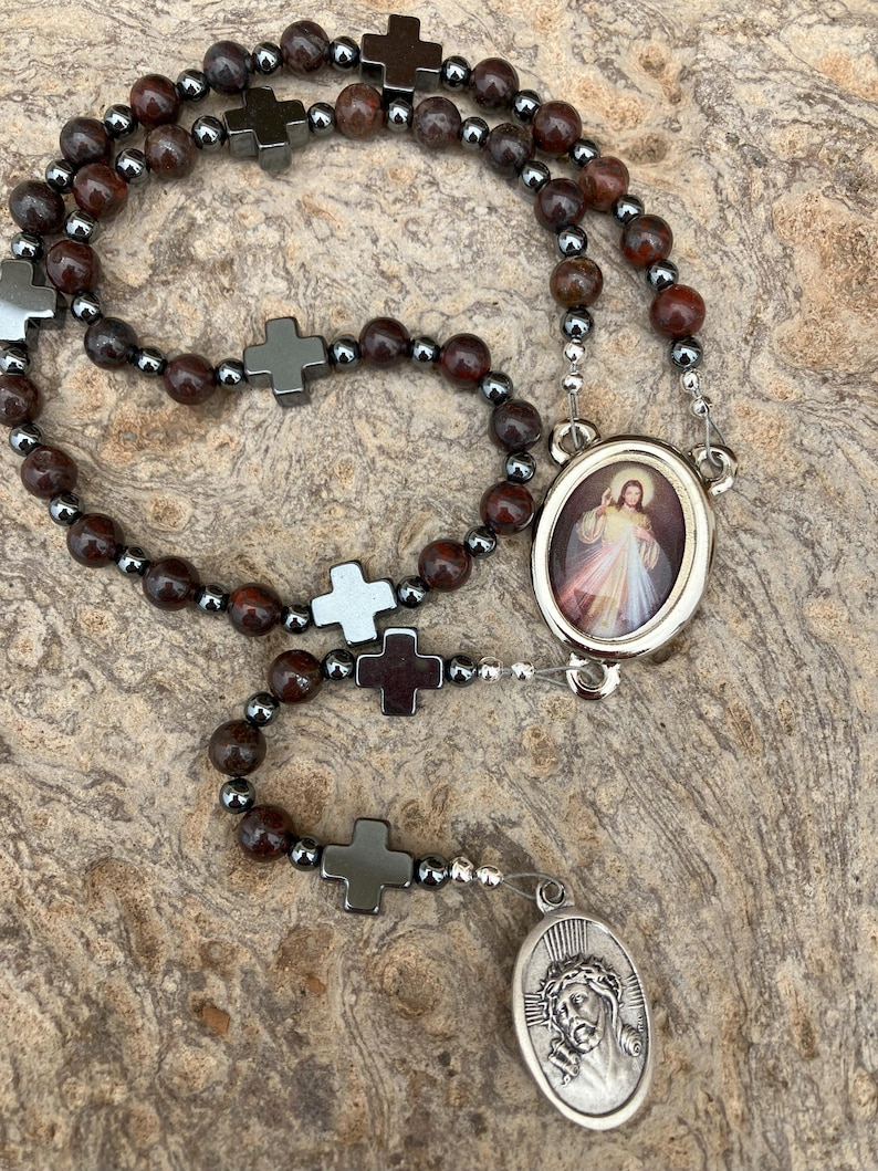 K&ouml;nnte beinhalten: Ein Rosenkranz mit dunkelroten und grauen Perlen, durchsetzt mit silberfarbenen Perlen und kreuzf&ouml;rmigen Akzenten. Der Rosenkranz hat zwei ovale Anh&auml;nger, einen mit einem Bild von Jesus und den anderen mit einer religi&ouml;sen Figur.