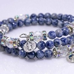 May include: A wrap bracelet featuring dark blue sodalite beads, interspersed with clear crystal accents and silver-tone metal spacers. The bracelet includes two small, round charms with religious imagery.