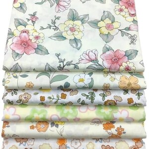 May include: A stack of floral fabric with various patterns. The top layer features large pink and yellow flowers with green leaves on a cream background. Other layers display different floral designs in shades of yellow, orange, and green.