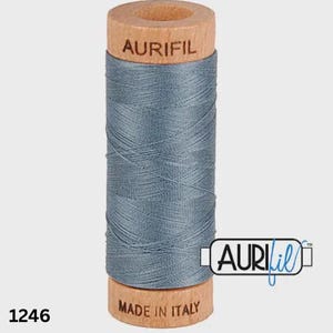 Puede incluir: Un carrete de hilo de coser azul grisáceo enrollado alrededor de un núcleo de madera. La parte superior del carrete de madera lleva el texto AURIFIL, mientras que la parte inferior dice MADE IN ITALY. El número 1246 está impreso en negro a la izquierda.