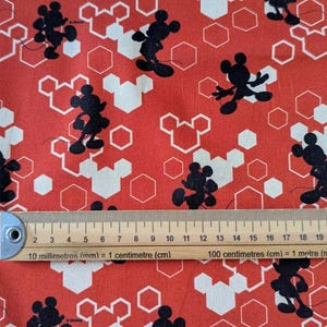Peut inclure: Tissu rouge avec un motif en nid d'abeille blanc et des silhouettes noires de Mickey Mouse. Le tissu est mesuré avec une règle en centimètres.
