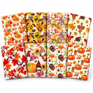 Könnte beinhalten: Acht gefaltete Stoffquadrate mit herbstlichen Mustern, darunter orangefarbene Kürbisse, gelbe Sonnenblumen, rote Ahornblätter, Truthähne und Zwerge auf weißem und cremefarbenem Hintergrund, ergänzt durch Motive wie Beeren, Äpfel und Birnen.