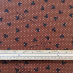Peut inclure: Tissu brun avec un motif répété de têtes de Mickey Mouse noires et le texte "mickey mouse" dans une police d'écriture cursive. Une règle est placée sur le tissu pour l'échelle.