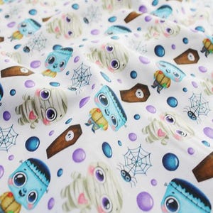 May include: White fabric with a repeating pattern of cartoon monsters, including mummies, Frankenstein's monsters, and coffins. The design also features spiderwebs, spiders, and purple and blue polka dots. The fabric is likely for Halloween-themed crafts.