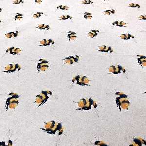 May include: A beige fabric with a repeating pattern of black and yellow bumblebees. The bumblebees are stylized and have a watercolor effect.