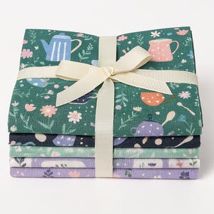 Könnte beinhalten: Ein Stapel aus sechs Stoffquadraten mit einem grünen Blumenmuster mit Teekannen und Tassen. Die Stoffquadrate sind mit einem cremefarbenen Band zusammengebunden.