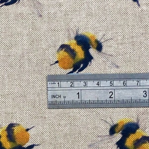 May include: Close-up of a beige fabric with a repeating pattern of yellow and black bumblebees. A ruler is placed on the fabric, showing measurements in inches.