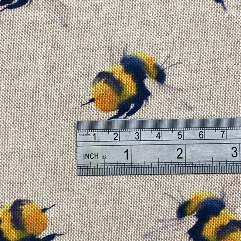 May include: Close-up of a beige fabric with a repeating pattern of yellow and black bumblebees. A ruler is placed on the fabric, showing measurements in inches.