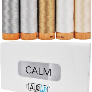 Puede incluir: Colección de cinco carretes de hilo en varios colores, incluyendo plata, dorado y blanco, con bases de madera. Encima, una caja blanca con la palabra "CALM" impresa.