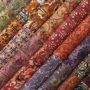 Op de afbeelding: Een verzameling stoffen rollen met diverse paisley- en bloempatronen. De stoffen hebben een reeks kleuren, waaronder tinten bruin, rood, paars en groen. De patronen bevatten ingewikkelde ontwerpen en gedetailleerde motieven, geschikt voor quilt- of knutselprojecten.