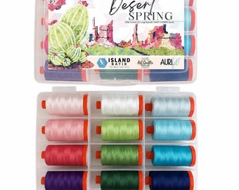 Könnte beinhalten: Ein durchsichtiger Kunststoff-Aufbewahrungskoffer mit zwölf großen Garnspulen in verschiedenen Farben, darunter Rosa, Grün, Blau und Violett. Das Etikett enthält den Text Desert Spring, 100% Cotton, 12 Large Spools, 50WT, Island Batik und Aurifil.