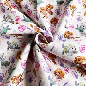 May include: White fabric with a repeating pattern of cartoon dogs from the Paw Patrol cartoon. The dogs are wearing their signature uniforms and are surrounded by paw prints, bones, and purple shields.