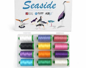 Könnte beinhalten: Eine weiße rechteckige Schachtel mit der Aufschrift Seaside, Vogelillustrationen und Logos, die einen durchsichtigen Kunststoff-Organizer mit zwölf großen Garnrollen aus 100% Baumwolle in verschiedenen Farben wie Weiß, Schwarz, Blau, Gelb und Pink enthält.