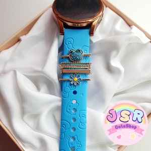 May include: A blue silicone watch strap with a Hello Kitty pattern. The strap has a gold-coloured metal charm with a blue gem and a gold-coloured metal charm with a white flower.