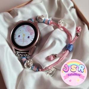 Könnte beinhalten: Eine roségoldene Smartwatch mit Hello Kitty-Zifferblatt, kombiniert mit einem rosa-blauen Kordelarmband. Die Uhr liegt auf weißem Stoff, mit einem dekorativen Charm am Armband. Das Zifferblatt zeigt "Hello Kitty 50th Anniversary".