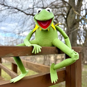 Puede incluir: Una marioneta de rana verde con una amplia sonrisa, cuello amarillo y ojos blancos está sentada en una valla de madera. Los brazos y las piernas de la rana están extendidos, y parece estar relajándose al aire libre. El fondo presenta una vista borrosa de árboles y cielo.