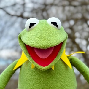 May include: A plush Kermit the Frog puppet with a green textured body, white eyes, and a red mouth. The puppet has yellow collar details and is smiling. The background is blurred, with tree branches visible.