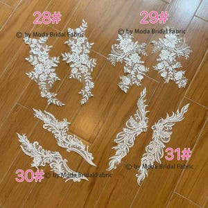 Può includere: Diverse applicazioni floreali in pizzo bianco sono esposte su una superficie di legno. Le applicazioni variano nel design, con alcuni motivi floreali dettagliati e altri che mostrano forme più intricate e fluide. I numeri 28#, 29#, 30# e 31# sono visibili.