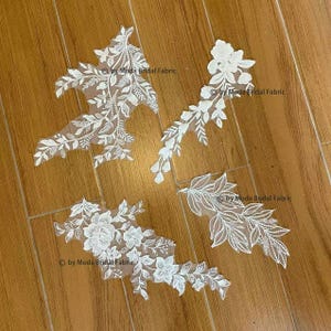 May include: Four white floral lace appliques with intricate leaf and flower designs. The appliques are on a light brown wooden surface. The text "by Moda Bridal Fabric" is visible on each applique.