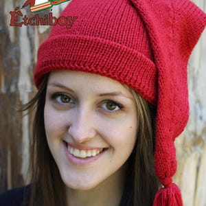 May include: A red knitted hat with a long, pointed, drooping earflap and a tassel. The hat is worn by a person with long brown hair.