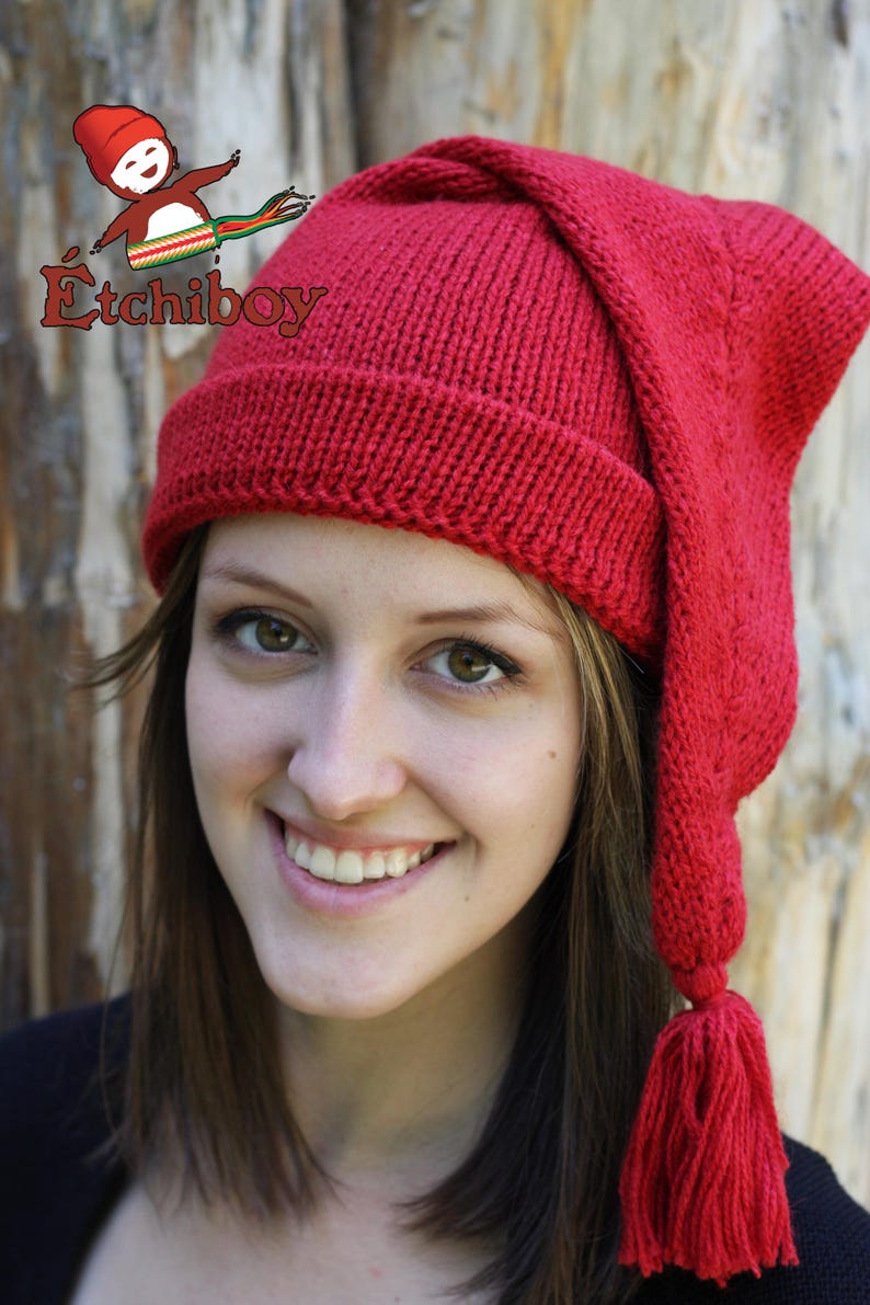 May include: A red knitted hat with a long, pointed, drooping earflap and a tassel. The hat is worn by a person with long brown hair.