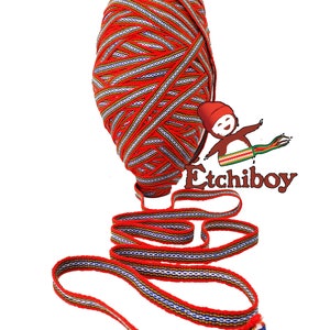 Peut inclure: Un ruban tissé rouge, blanc, bleu et or avec un motif répétitif. Le ruban est enroulé en boule et une longueur du ruban est déroulée.