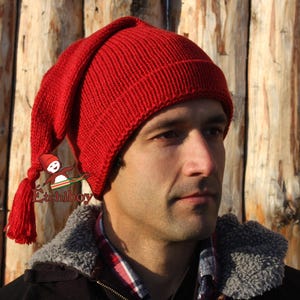 May include: A red knitted hat with a long pointed top and a tassel. The hat is worn by a person with a beard and a brown jacket.