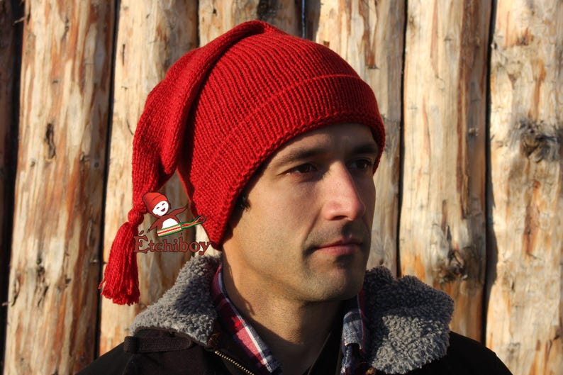 May include: A red knitted hat with a long pointed top and a tassel. The hat is worn by a person with a beard and a brown jacket.