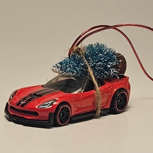 May include: A red Corvette sports car with a small blue Christmas tree tied to the roof. The car is a miniature ornament.